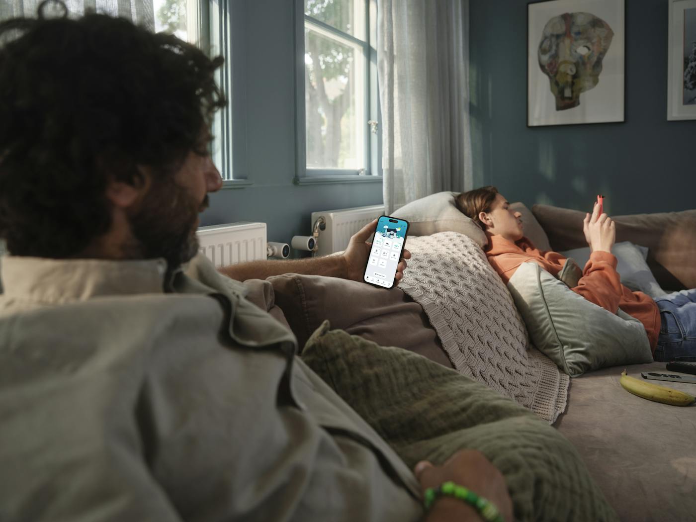 To personer slapper av i en sofa, den ene ser på strømforbruket i Tibber-appen på mobilen